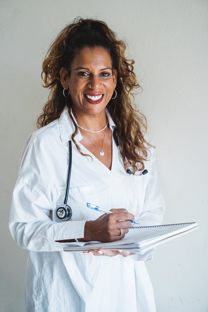Portrait of a smiling female doctor holding a notebook, wearing a white coat and stethoscope, against a plain background.